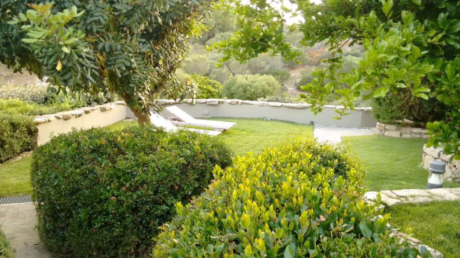 Lush green garden featuring manicured hedges, vibrant shrubs, and a serene landscape with a pool in the background, ideal for outdoor relaxation. | Only on SopranoVillas