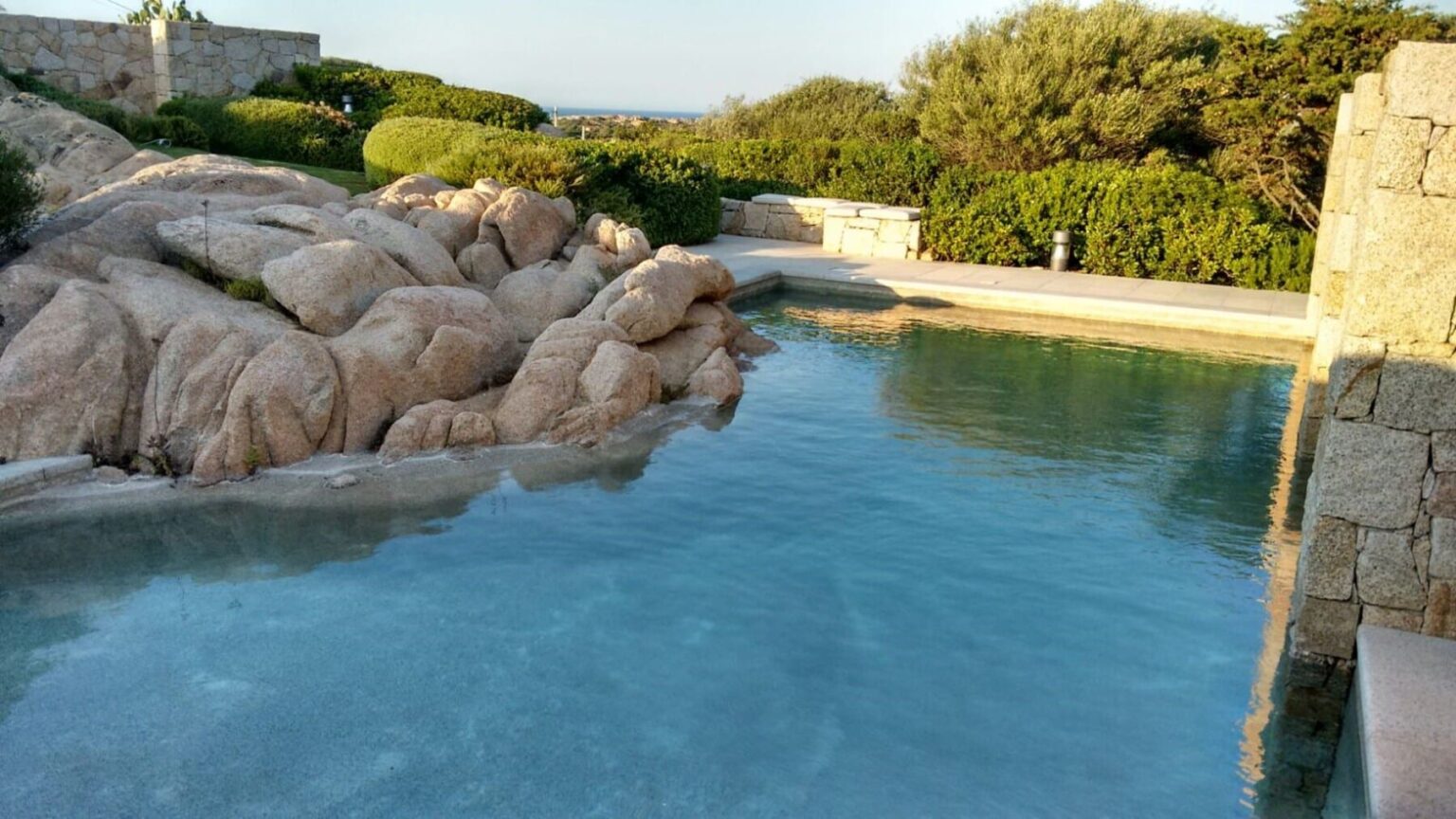 A serene outdoor swimming pool surrounded by natural rock formations and lush greenery, with clear blue water reflecting the sky. Ideal for relaxation and enjoying nature. | Only on SopranoVillas