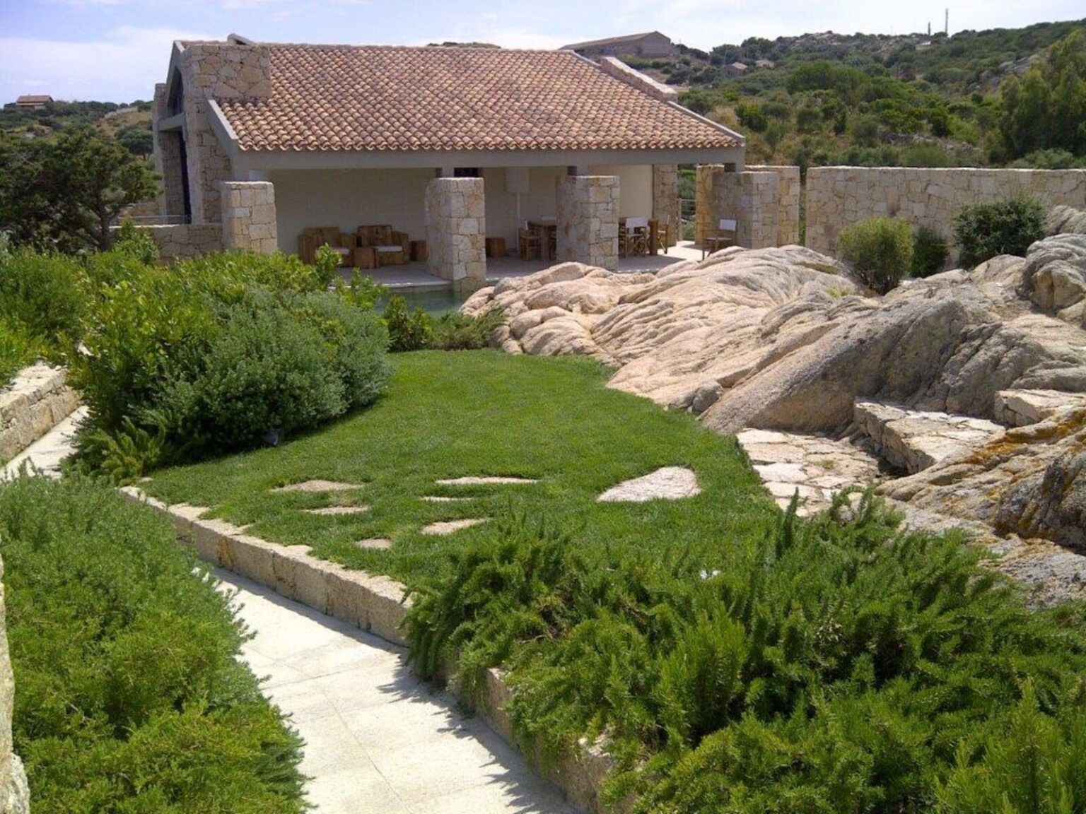 A serene landscape featuring a modern house with a tiled roof, surrounded by lush greenery and rocky terrain, showcasing a well-maintained garden path leading to the entrance. | Only on SopranoVillas