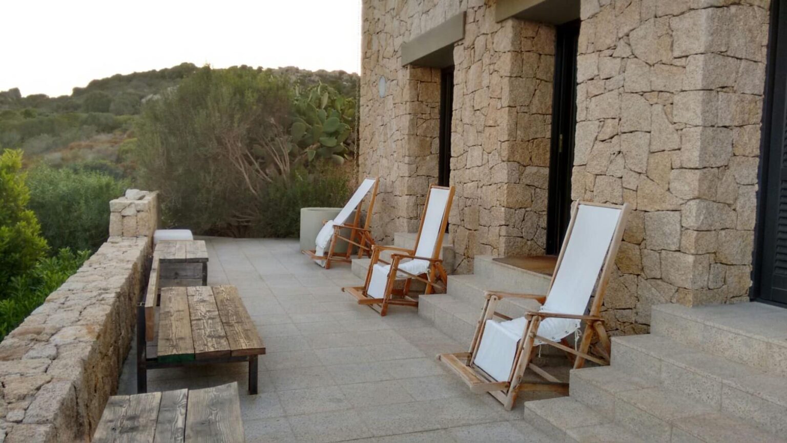 Cozy outdoor seating area with wooden lounge chairs and a stone wall, surrounded by greenery, ideal for relaxation and enjoying nature. | Only on SopranoVillas