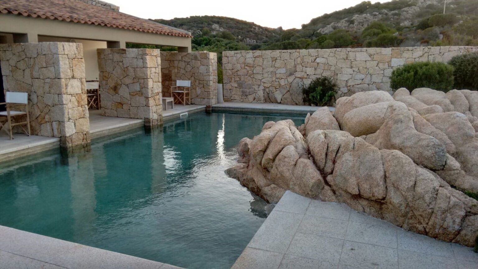 Luxury outdoor pool surrounded by natural stone features and a serene landscape, ideal for relaxation and leisure. The setting includes a rustic stone wall and comfortable seating, perfect for a tranquil retreat. | Only on SopranoVillas