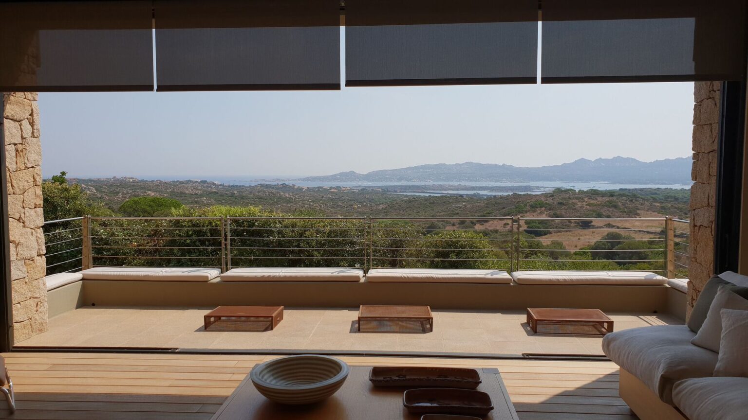 Modern outdoor living space with a panoramic view of lush green hills and a serene lake, featuring a comfortable seating area and natural stone details. | Only on SopranoVillas