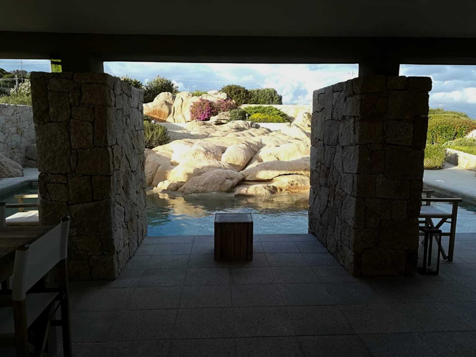 View from a modern stone archway overlooking a serene pool and landscaped garden with natural rock formations and colorful flowers. | Only on SopranoVillas