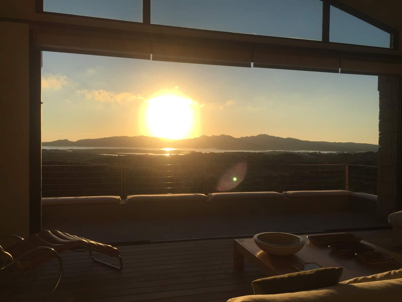 Sunset view from a modern living room with large windows, showcasing mountains and a tranquil lake in the distance. The scene features a comfortable seating area and natural light illuminating the space, creating a serene atmosphere. | Only on SopranoVillas