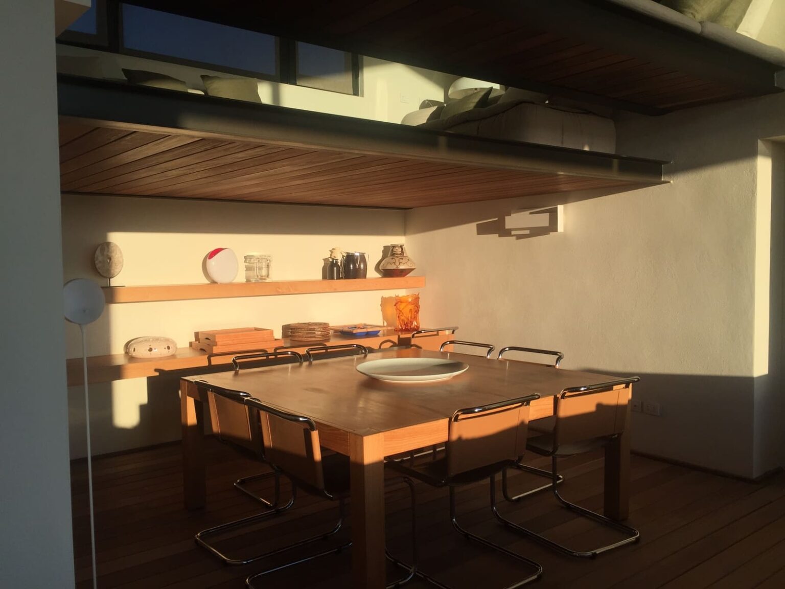 Modern kitchen interior featuring a wooden dining table with chairs, illuminated by natural sunlight. Shelves display decorative items and kitchenware, creating a warm and inviting atmosphere. | Only on SopranoVillas