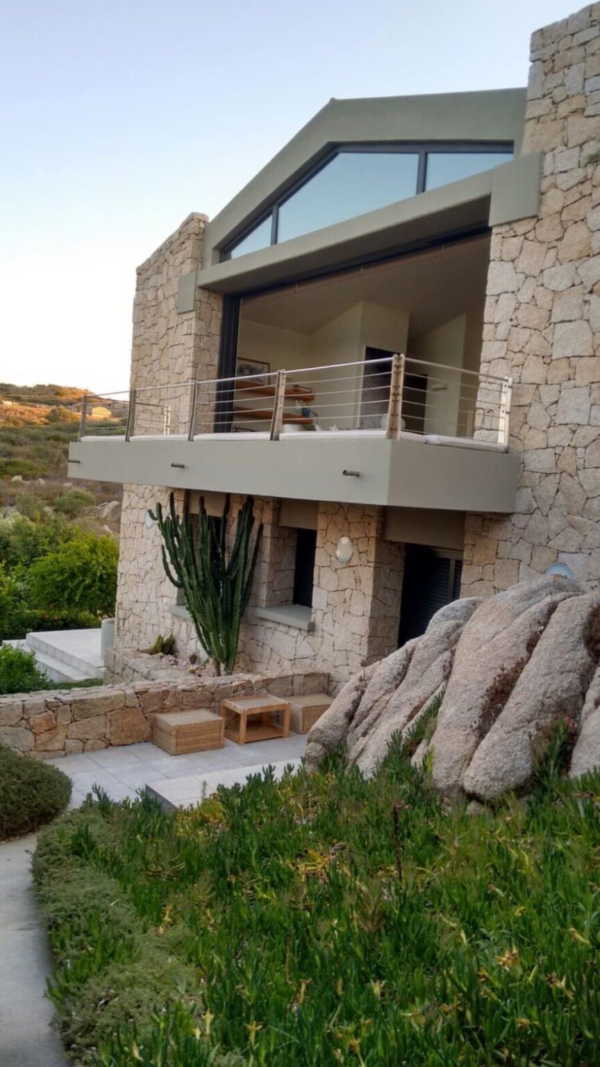 Modern stone house with a balcony, featuring a tall cactus and large rocks in the foreground, set against a natural landscape. | Only on SopranoVillas