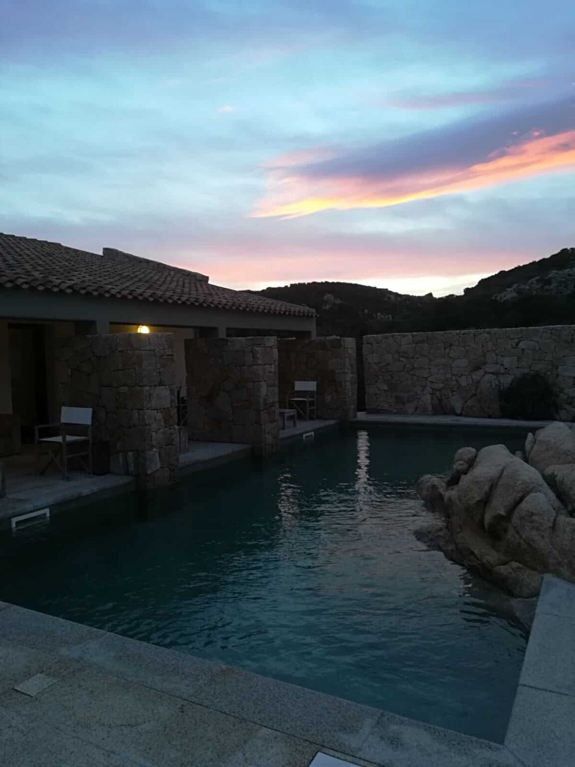 Serene evening view of a stone-lined swimming pool surrounded by a rustic villa, under a colorful sunset sky with hues of pink and blue. | Only on SopranoVillas