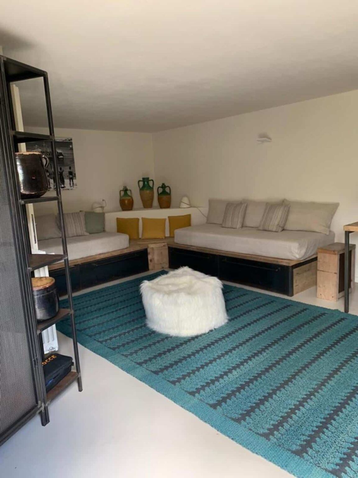 Cozy living room featuring two daybeds with decorative pillows, a fluffy white pouf, and a textured teal area rug. The space is complemented by rustic decor, including green and brown pottery, and a modern shelving unit. | Only on SopranoVillas