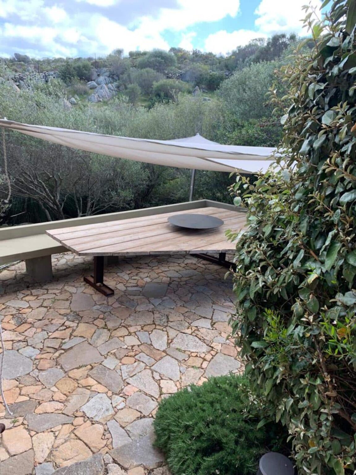 Outdoor dining area featuring a wooden table with a circular centerpiece, surrounded by greenery and shaded by a fabric canopy. Stone flooring adds a rustic touch to the serene setting. | Only on SopranoVillas