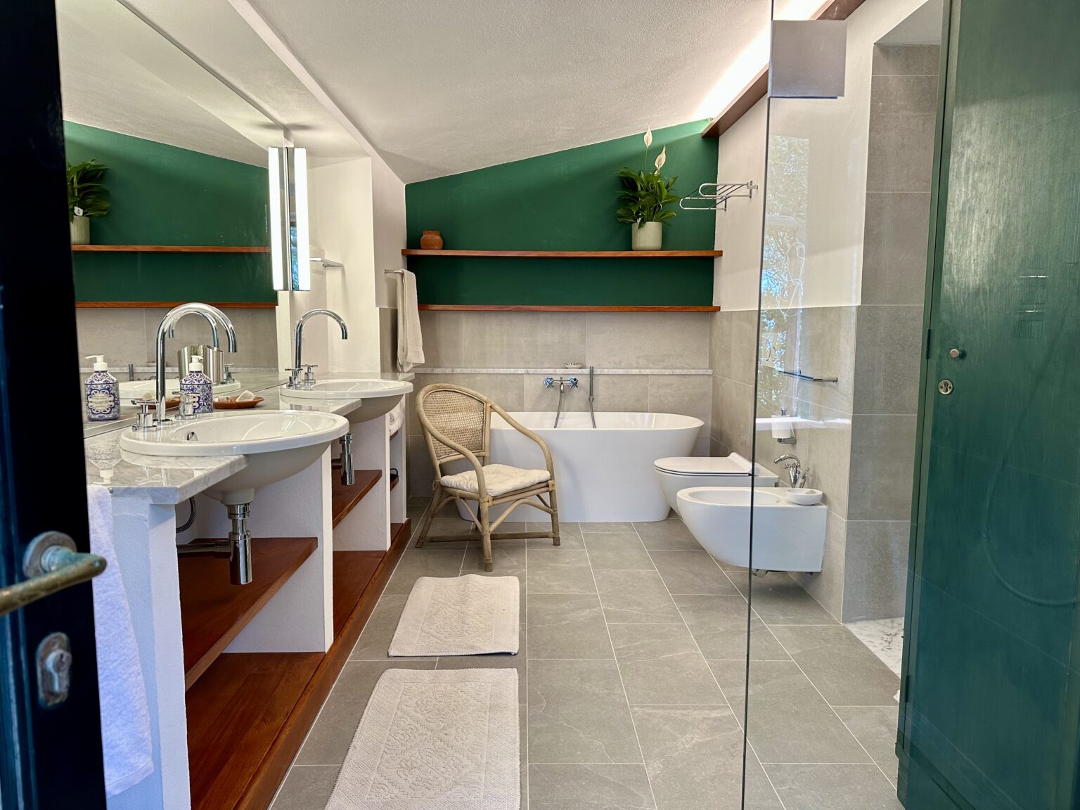 Modern bathroom design featuring dual sinks, a freestanding bathtub, and a bidet, complemented by green accent walls and wooden shelves. The space includes contemporary lighting and natural elements, creating a stylish and functional atmosphere. | Only on SopranoVillas