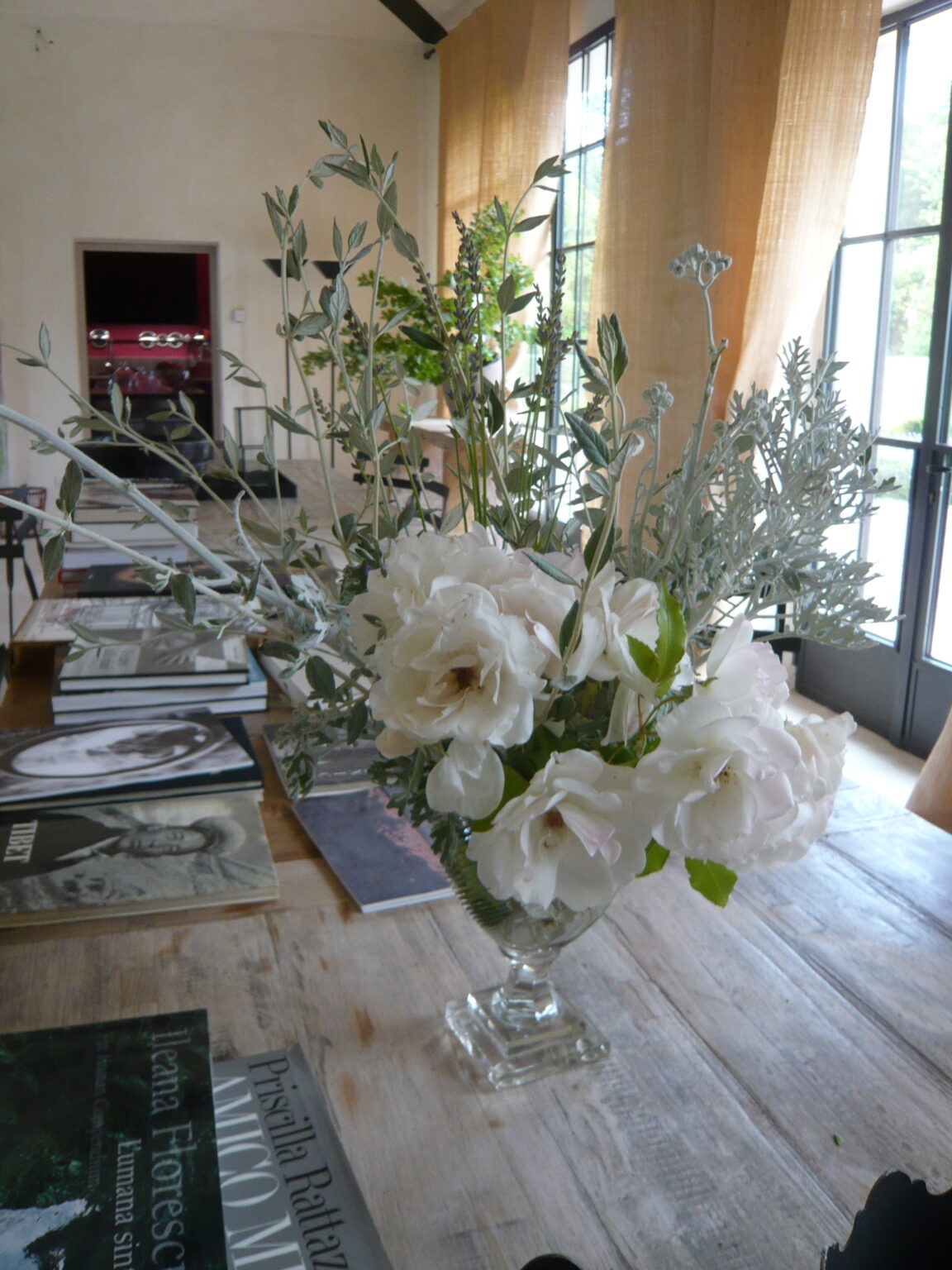 A beautiful floral arrangement featuring soft white roses and greenery in a crystal vase, placed on a wooden table surrounded by art books in a bright, modern interior with large windows and natural light. | Only on SopranoVillas