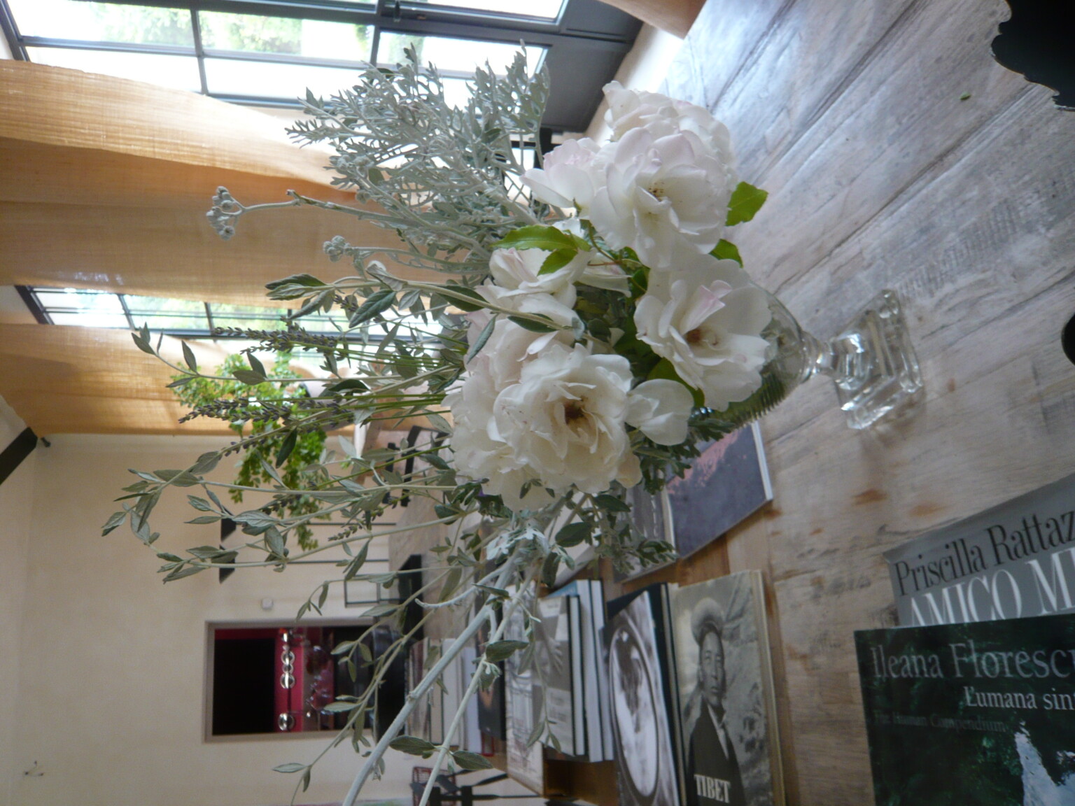 A beautiful floral arrangement featuring soft white roses and greenery in a crystal vase, placed on a wooden table surrounded by art books in a bright, modern interior with large windows and natural light. | Only on SopranoVillas