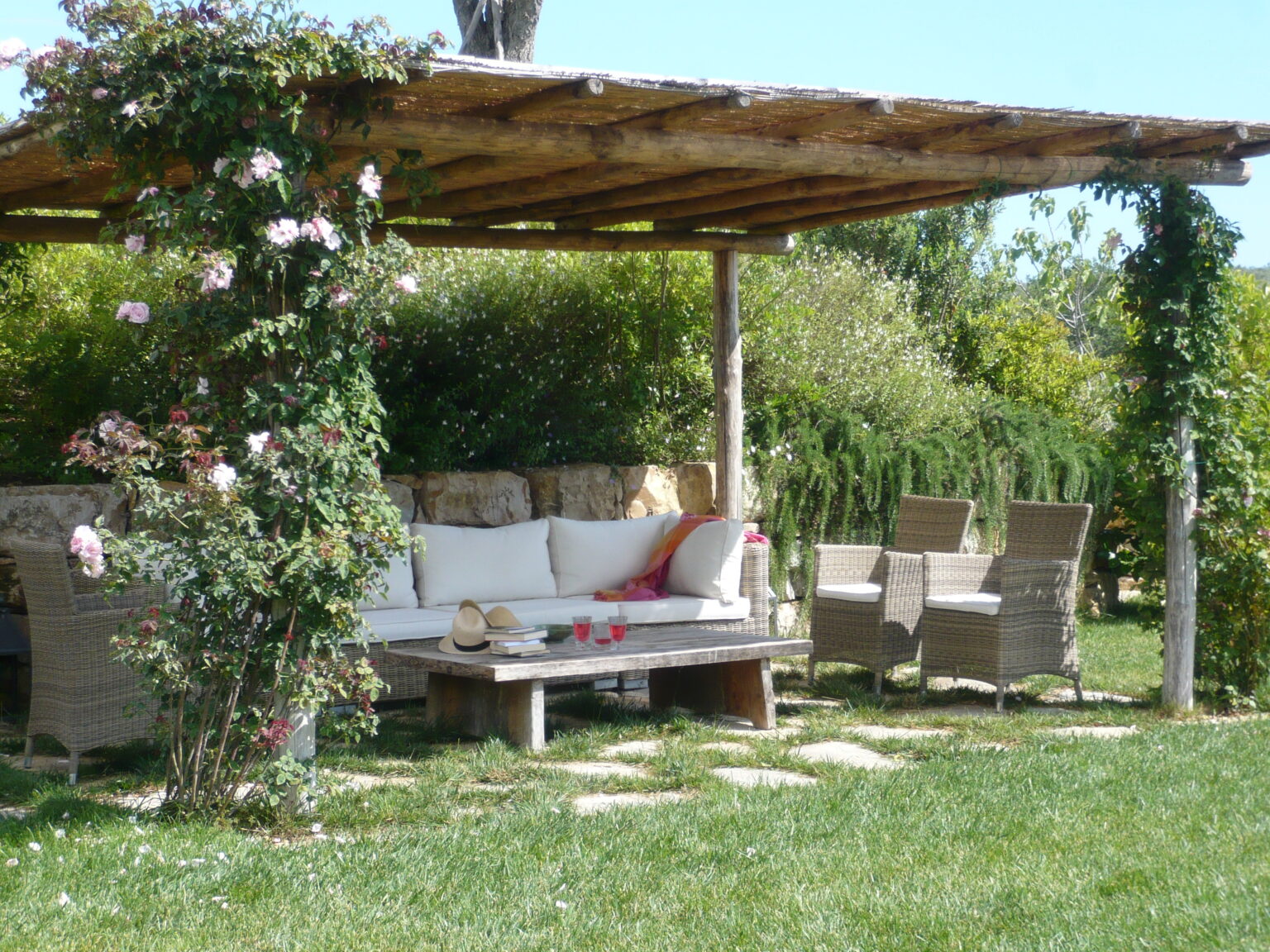 Cozy outdoor seating area with a wooden pergola adorned with climbing roses and greenery, featuring a comfortable sofa and a coffee table surrounded by wicker chairs, set in a lush garden. | Only on SopranoVillas