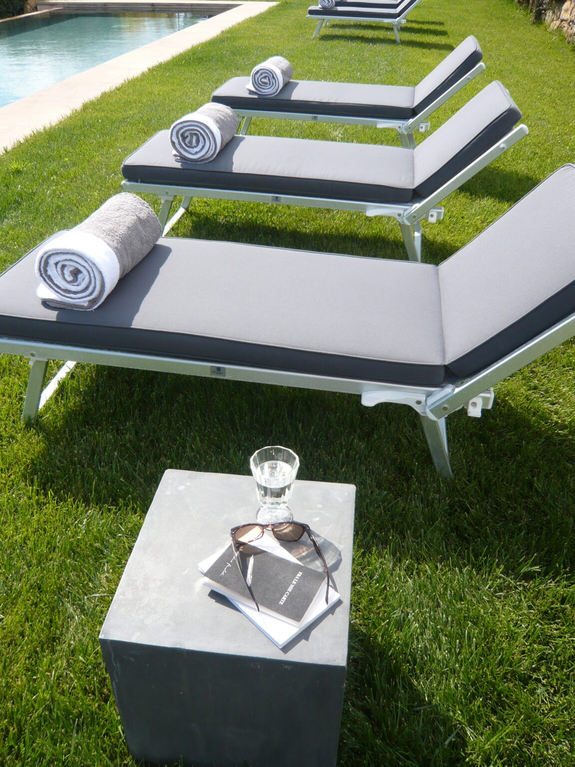 Lounge chairs with rolled towels beside a swimming pool on a sunny day, featuring a concrete side table with sunglasses, a glass of water, and a notebook. | Only on SopranoVillas