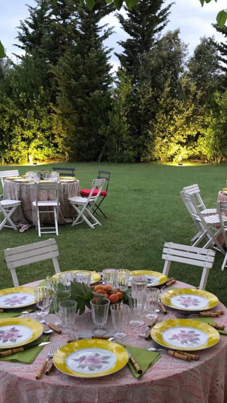 Elegant outdoor dining setup featuring a beautifully arranged table with floral plates, crystal glassware, and fresh fruit centerpiece. Surrounded by lush greenery and illuminated by soft lighting, this scene is perfect for a summer gathering or special event. | Only on SopranoVillas