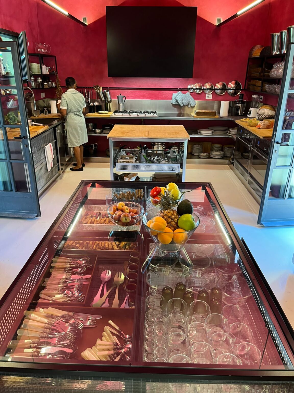 Modern kitchen interior featuring a chef preparing food, with a vibrant red wall, stainless steel appliances, and a central island. The glass display showcases an organized array of cutlery, glasses, and fresh fruits, creating an inviting culinary atmosphere. | Only on SopranoVillas