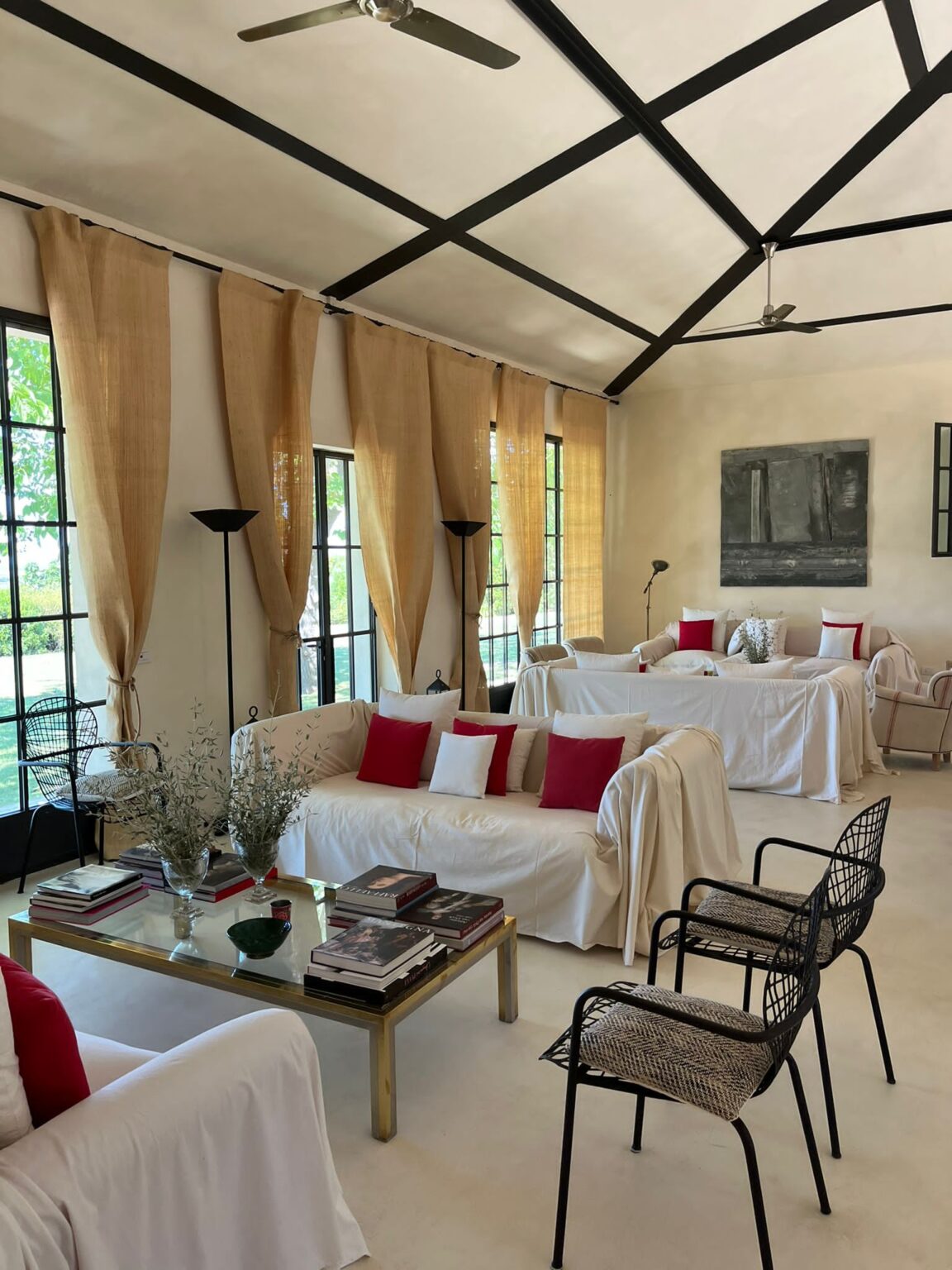 Bright and airy living room featuring white sofas with red and white cushions, a glass coffee table stacked with books, and elegant black chairs. Natural light streams through large windows adorned with sheer curtains, enhancing the modern decor with a minimalist aesthetic. | Only on SopranoVillas