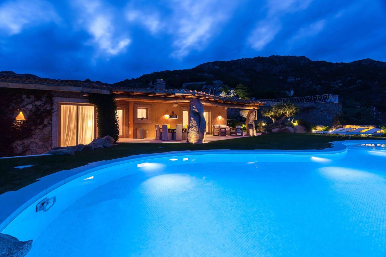 Luxurious villa with a glowing pool at dusk, surrounded by lush greenery and rocky hills, featuring a cozy outdoor dining area and warm ambient lighting. Ideal for vacation rentals or serene getaways. | Only on SopranoVillas
