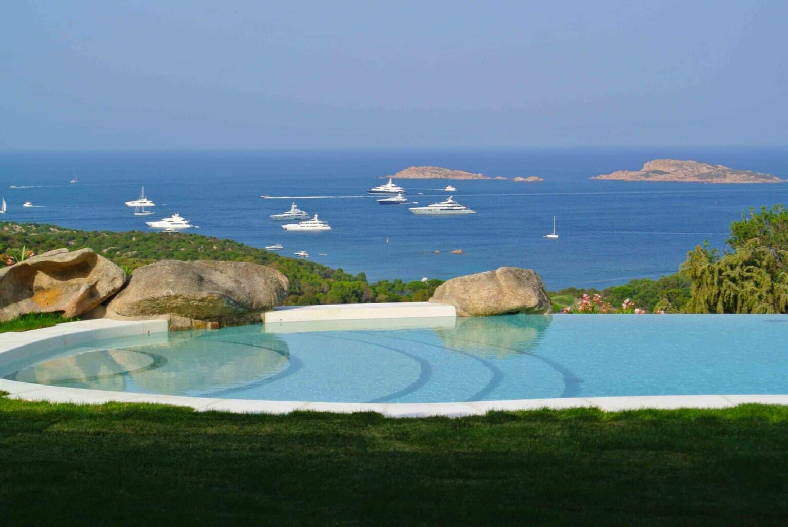 Luxurious infinity pool overlooking a serene blue sea dotted with yachts and rocky islands, surrounded by lush greenery and natural stone formations. | Only on SopranoVillas