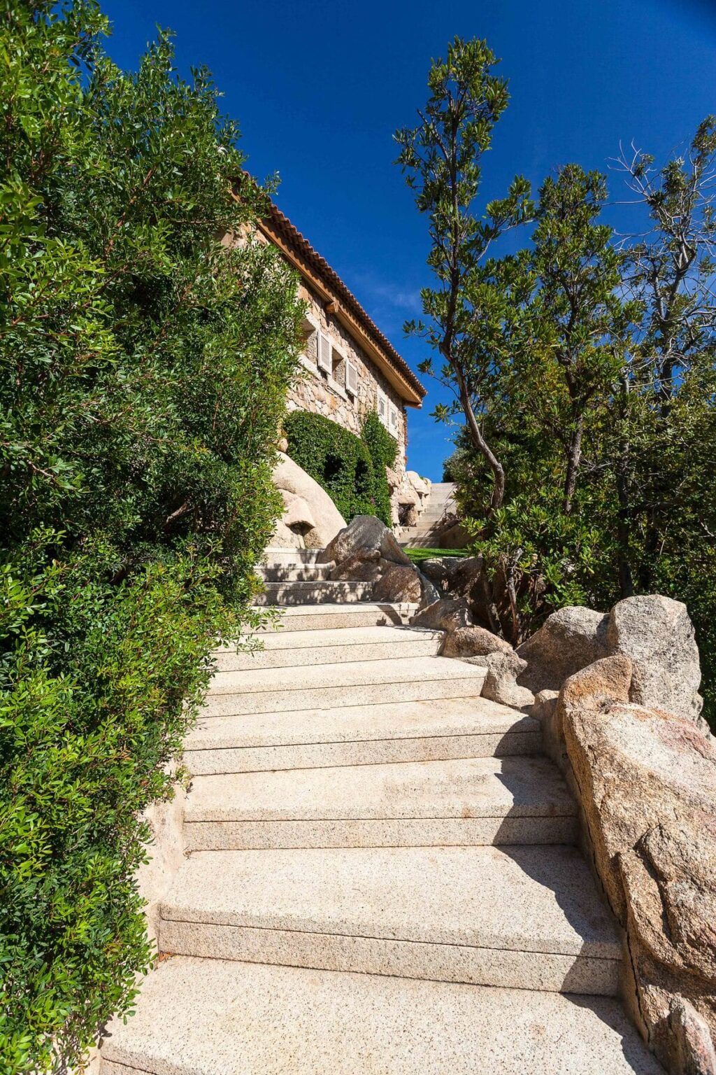 Stone staircase leading to a beautiful house surrounded by lush greenery and large rocks, under a clear blue sky. | Only on SopranoVillas