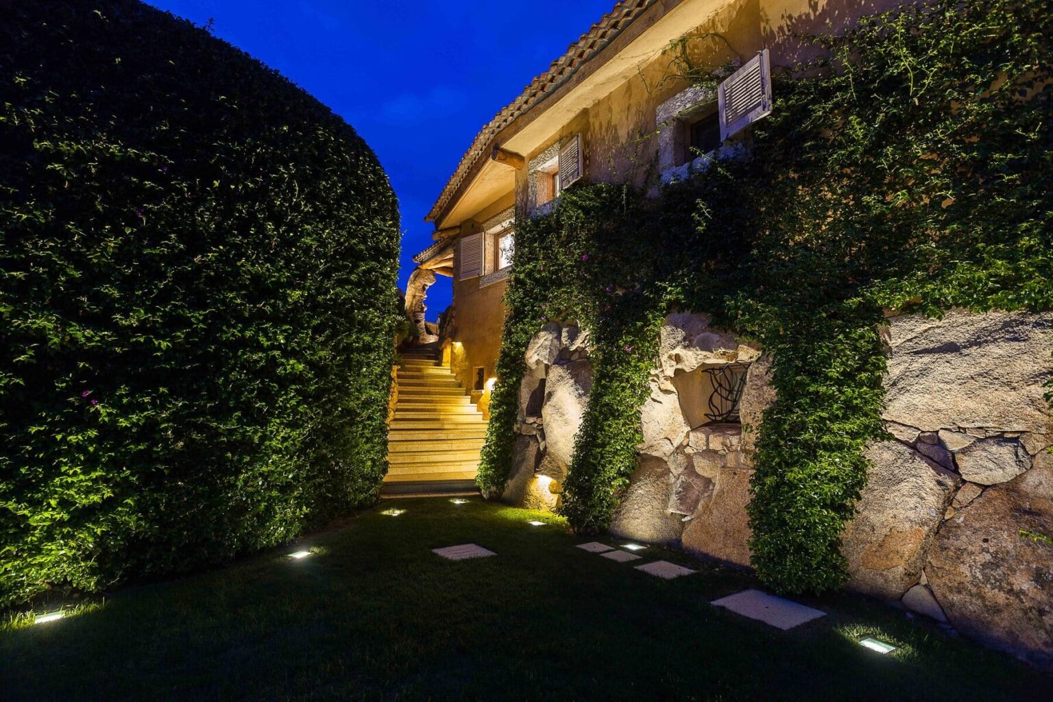 Pathway leading to a well-lit staircase beside a beautifully landscaped garden at dusk, featuring lush greenery and stone accents. | Only on SopranoVillas