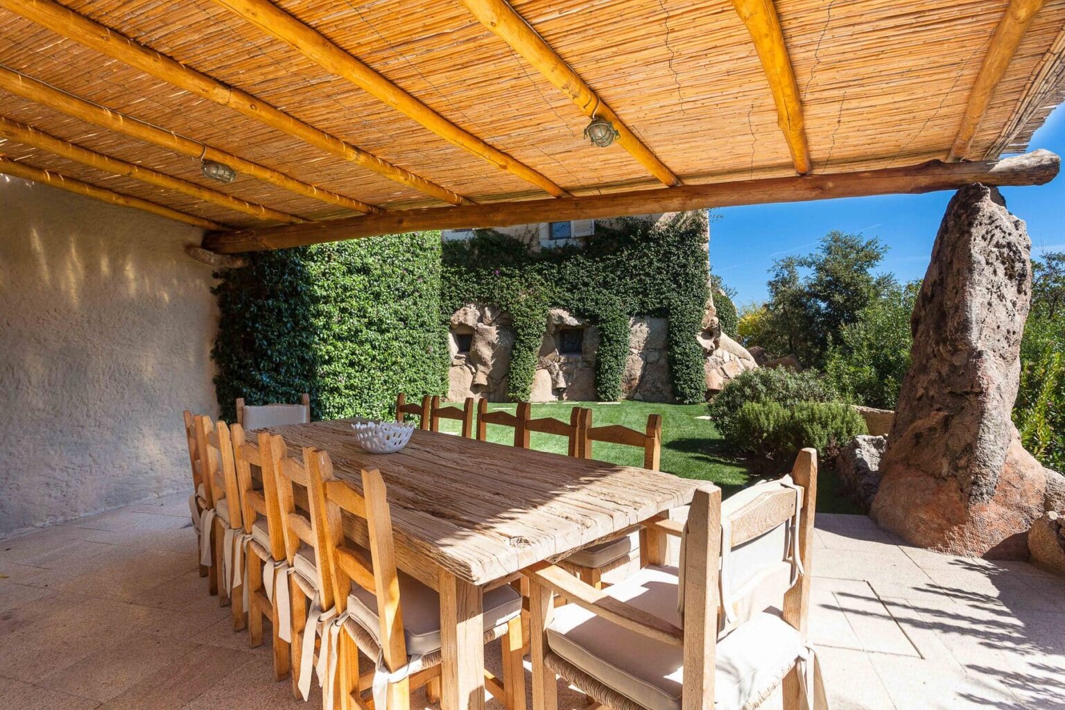 Outdoor dining area featuring a large wooden table surrounded by chairs, set under a bamboo roof. Lush greenery and rock formations create a natural backdrop, ideal for al fresco meals. | Only on SopranoVillas