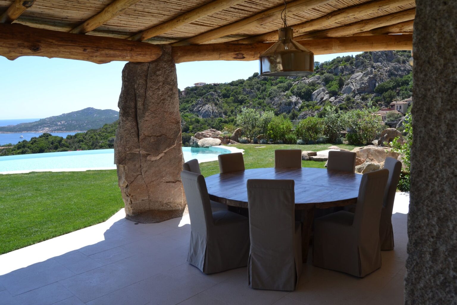 Outdoor dining area with a round table and upholstered chairs, surrounded by lush greenery and a scenic view of a pool and hills. Ideal for relaxation and entertaining in a tranquil setting. | Only on SopranoVillas