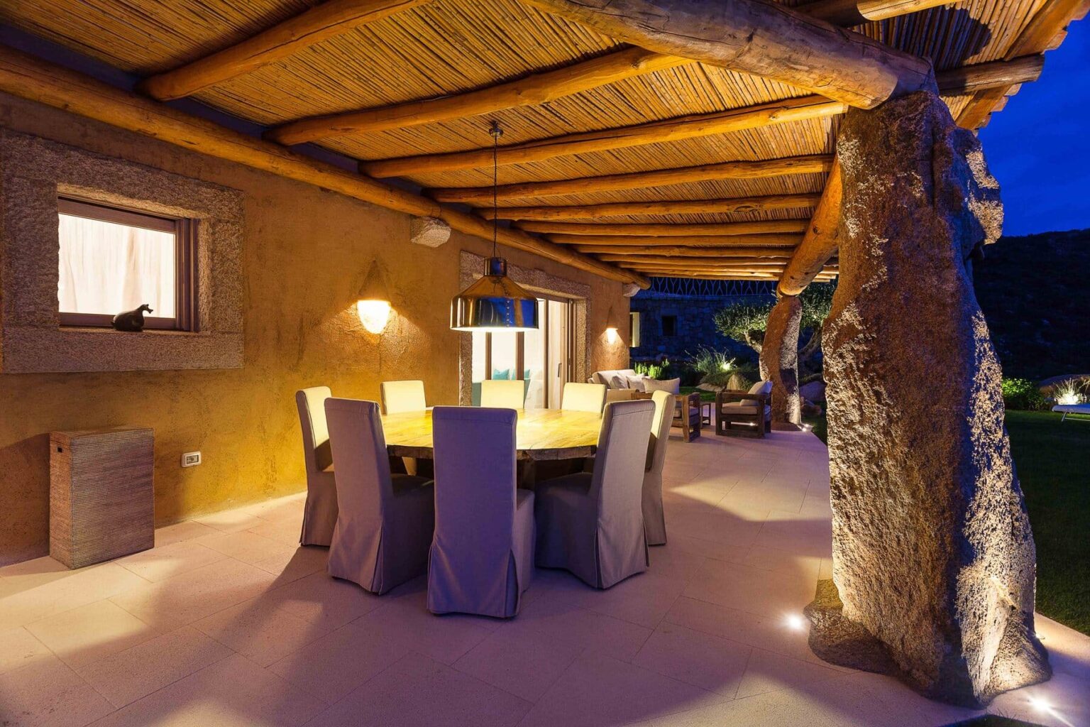 Outdoor dining area with a rustic wooden ceiling, stone pillars, and elegant table settings, illuminated at night in a serene setting. | Only on SopranoVillas