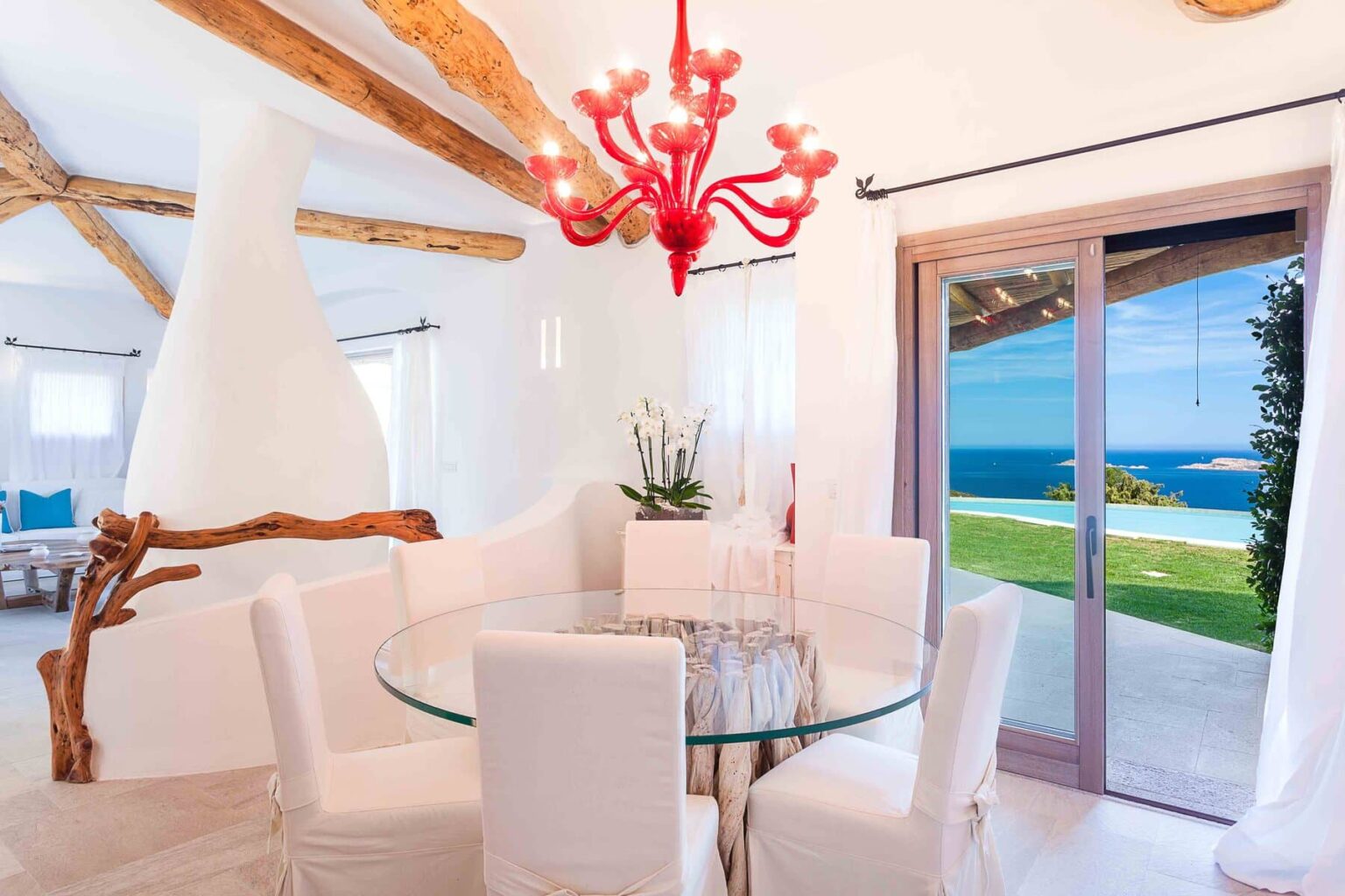 Modern dining area with a glass table and white chairs, featuring a coral chandelier and a scenic ocean view through large sliding doors, set in a bright, airy room with wooden beams. | Only on SopranoVillas