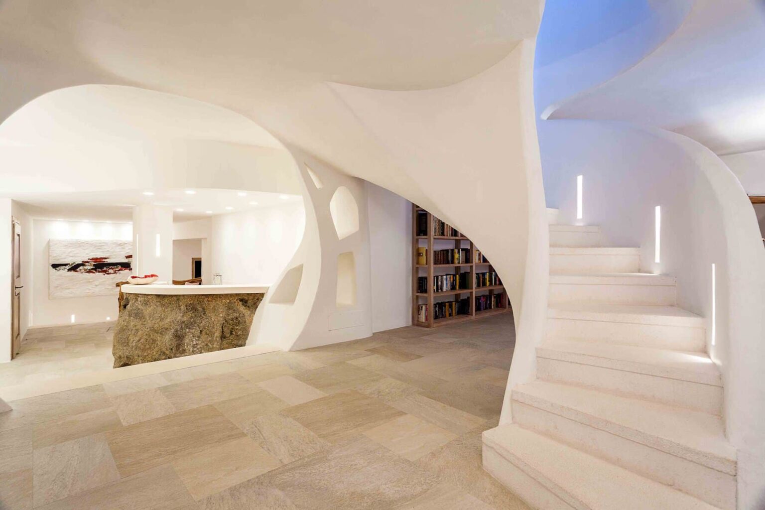 Modern interior design featuring a spacious foyer with sculptural white walls and elegant curved staircase. The area includes a natural stone bar and a bookshelf, complemented by soft lighting and contemporary artwork. Ideal for showcasing innovative architectural elements in residential spaces. | Only on SopranoVillas