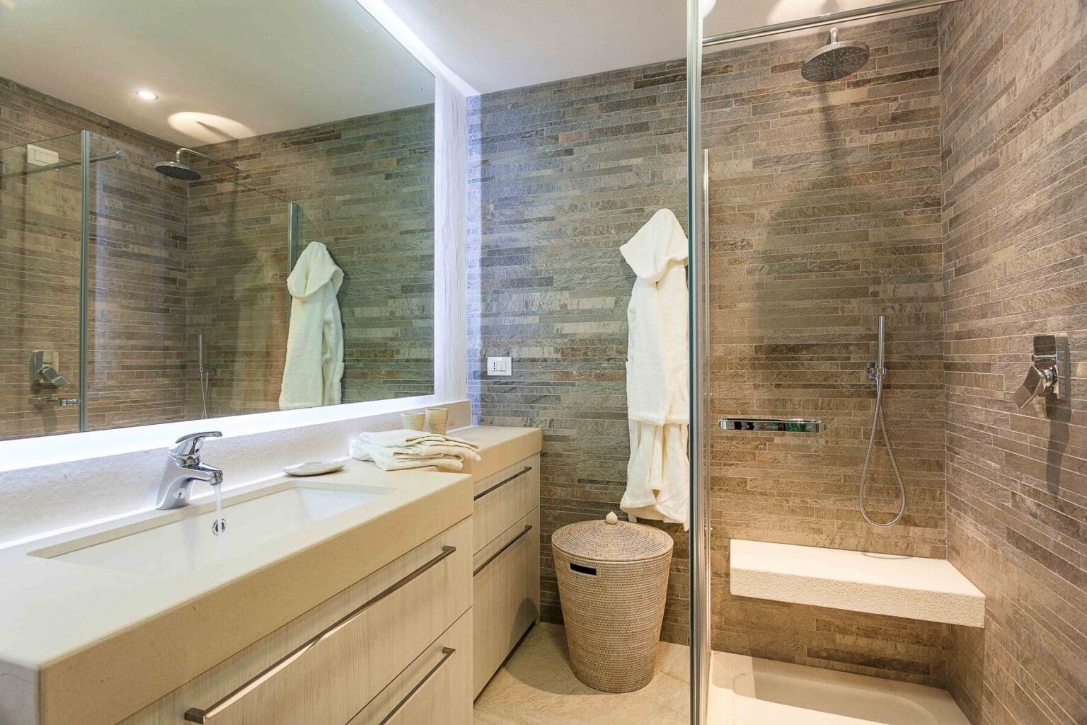 Modern bathroom design featuring a sleek vanity with dual sinks, a large mirror, and a glass shower. The walls are adorned with textured stone tiles, complemented by a minimalist towel rack and a stylish wicker basket. Soft lighting enhances the contemporary aesthetic. | Only on SopranoVillas
