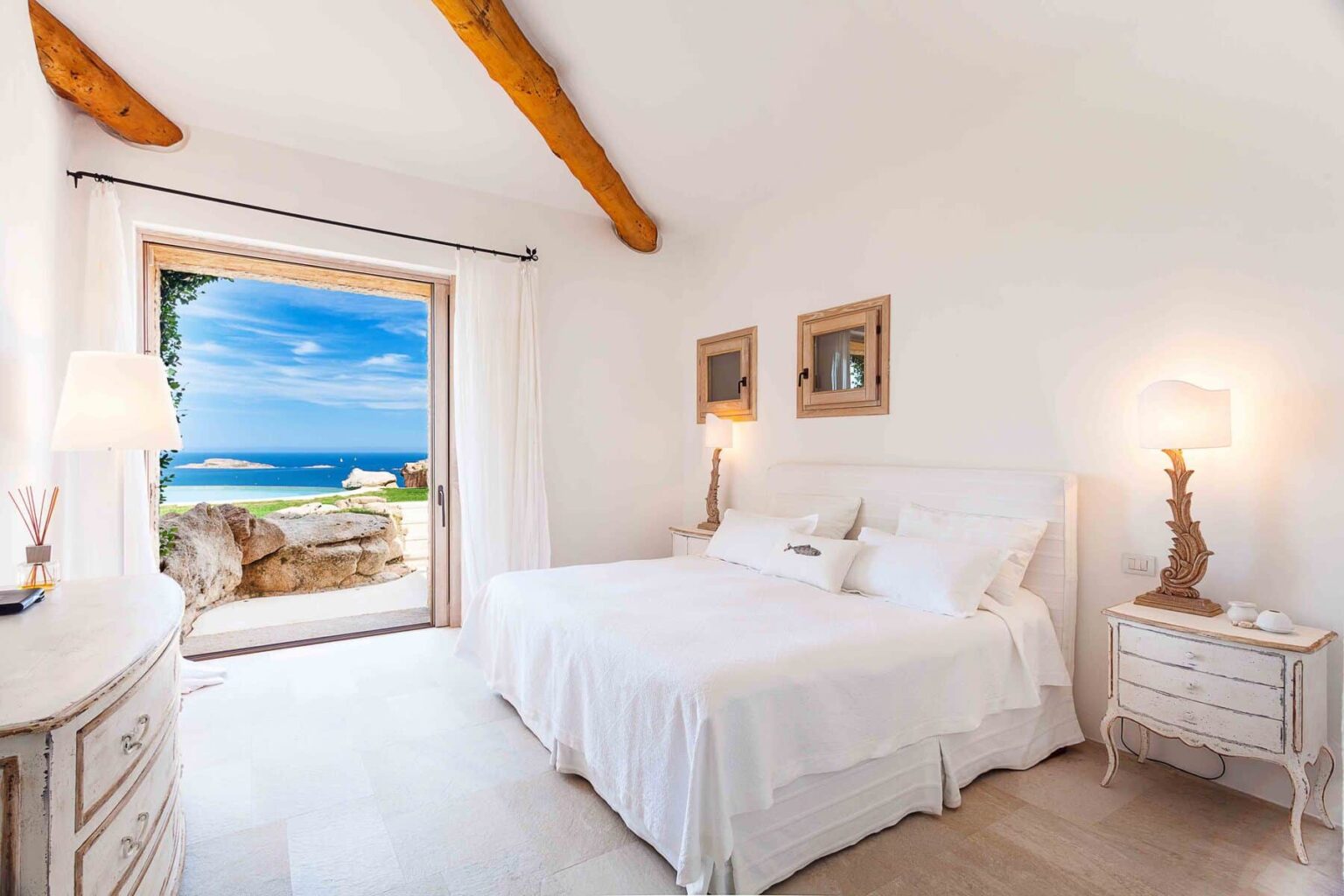 Bright and airy bedroom with a white bedspread, featuring large windows that open to a stunning ocean view. The room includes stylish decor with wooden beams and elegant lighting, creating a serene coastal retreat atmosphere. | Only on SopranoVillas