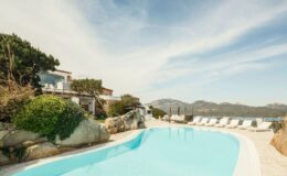 Luxurious villa with a pristine swimming pool, surrounded by lush greenery and rocky landscapes, offering stunning mountain and sea views in a tranquil setting.