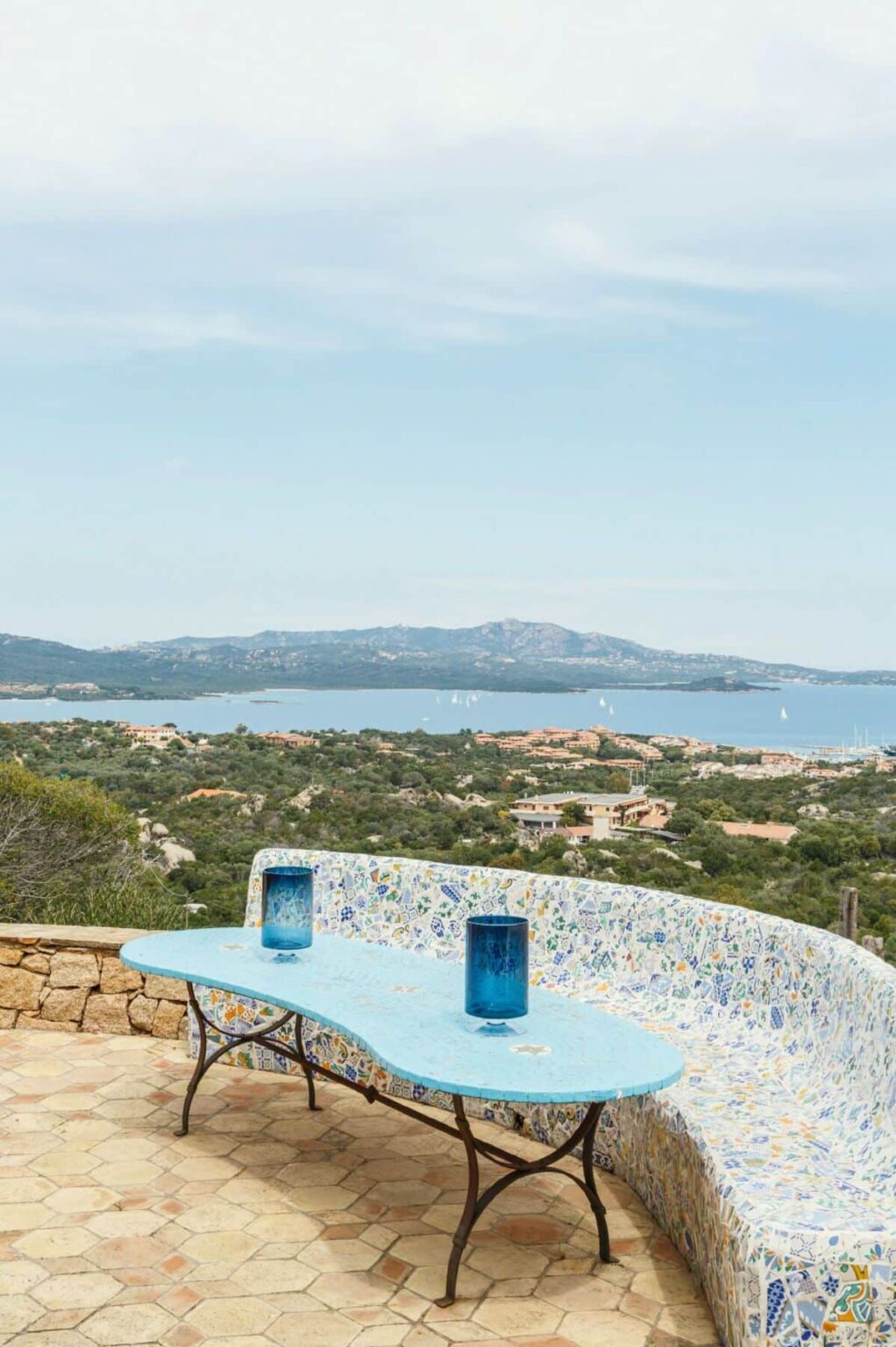 Scenic view of a coastal landscape with a blue sea and distant hills, featuring lush greenery in the foreground and a mosaic-patterned wall in the bottom corner. Ideal for travel and nature enthusiasts. | Only on SopranoVillas