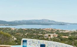 Scenic view of a coastal landscape with a blue sea and distant hills, featuring lush greenery in the foreground and a mosaic-patterned wall in the bottom corner. Ideal for travel and nature enthusiasts.