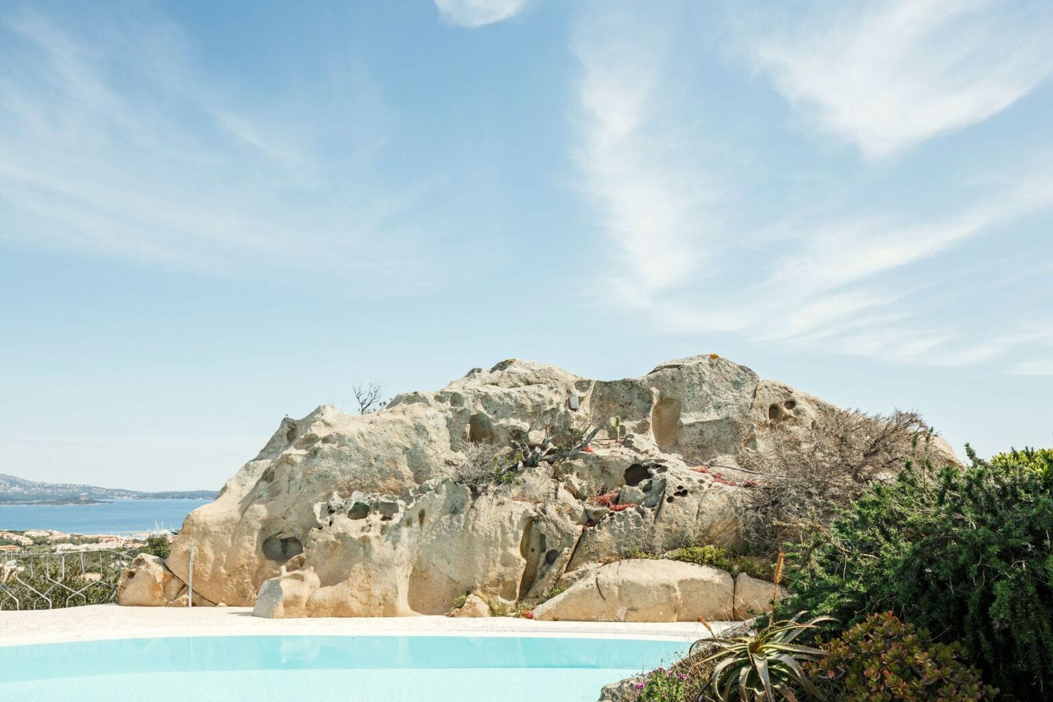 Scenic view of a large rock formation overlooking a tranquil swimming pool, with a coastal landscape and clear blue sky in the background, highlighting natural beauty and outdoor living. | Only on SopranoVillas