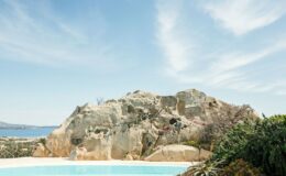 Scenic view of a large rock formation overlooking a tranquil swimming pool, with a coastal landscape and clear blue sky in the background, highlighting natural beauty and outdoor living.