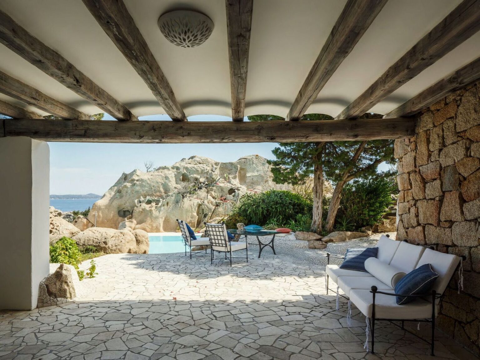 Cozy outdoor patio with stone walls and wooden beams, featuring a comfortable sofa and dining table, overlooking a serene pool and rocky coastline. Perfect for relaxation and enjoying coastal views. | Only on SopranoVillas