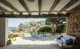 Cozy outdoor patio with stone walls and wooden beams, featuring a comfortable sofa and dining table, overlooking a serene pool and rocky coastline. Perfect for relaxation and enjoying coastal views.