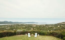 Scenic view of a coastal landscape with two lounge chairs in the foreground, overlooking greenery and a serene ocean in the distance under a cloudy sky.