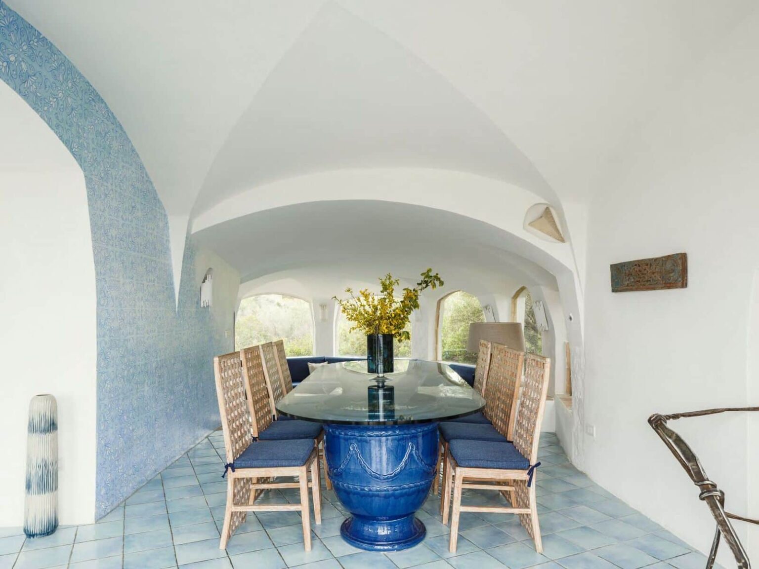 Bright and airy dining area featuring a large glass table surrounded by wooden chairs, with a blue vase filled with flowers as the centerpiece. The walls are painted white with blue accents, and the ceiling showcases an elegant arch design, creating a modern and inviting atmosphere. | Only on SopranoVillas