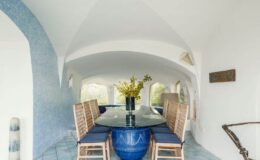 Bright and airy dining area featuring a large glass table surrounded by wooden chairs, with a blue vase filled with flowers as the centerpiece. The walls are painted white with blue accents, and the ceiling showcases an elegant arch design, creating a modern and inviting atmosphere.