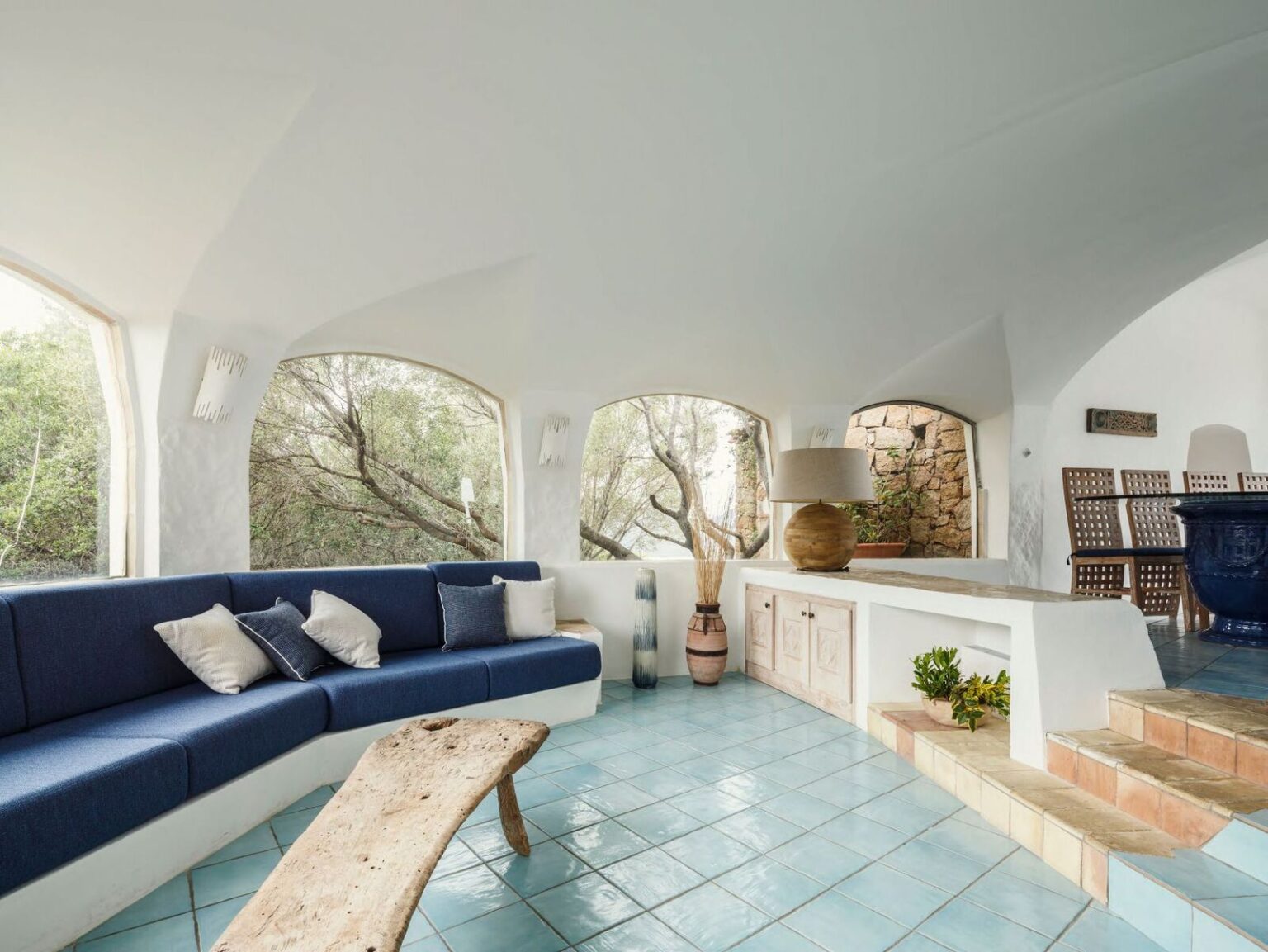 Spacious living room featuring a curved blue sofa with decorative pillows, a rustic wooden coffee table, and large windows showcasing a natural landscape. The light-colored walls and tiled floor create a bright, airy atmosphere, complemented by unique decor elements like a lamp and potted plants. | Only on SopranoVillas