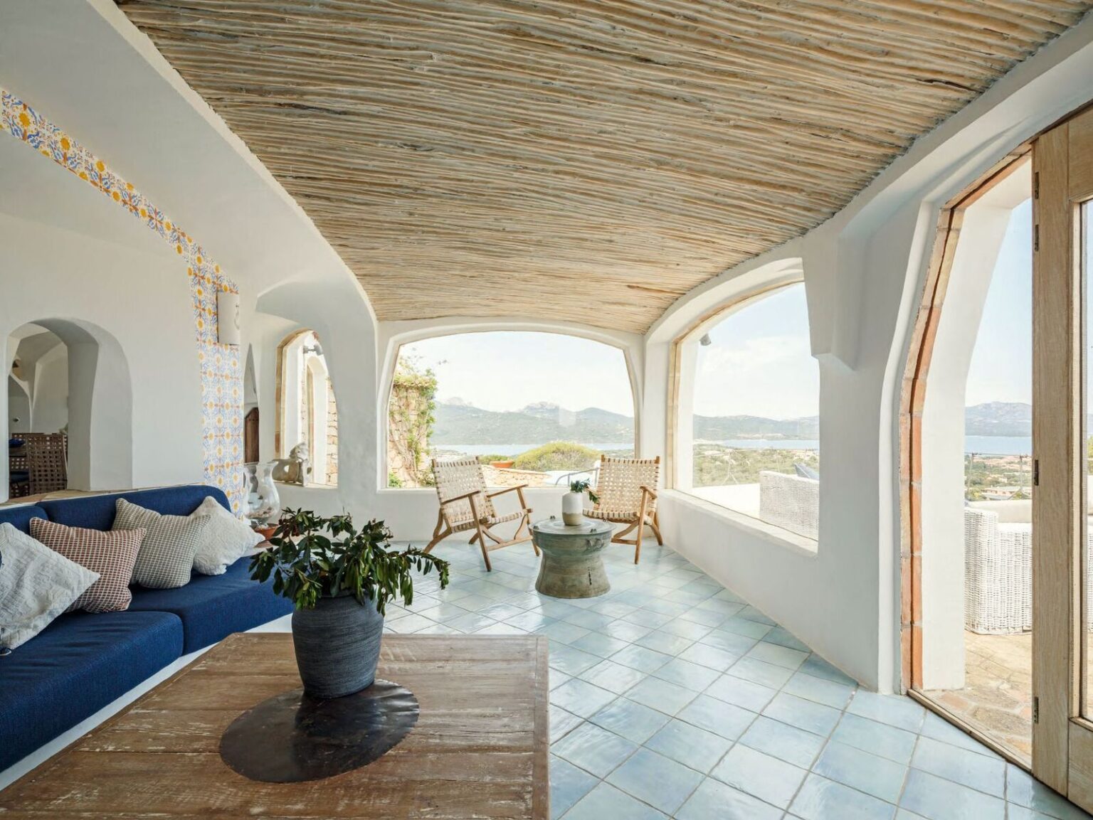 Cozy and modern living room with a blue sofa, wooden furniture, and large windows showcasing a scenic view of mountains and a serene landscape. The space features a natural straw ceiling and stylish decor, creating a relaxing atmosphere. | Only on SopranoVillas