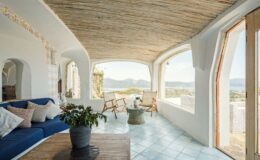 Cozy and modern living room with a blue sofa, wooden furniture, and large windows showcasing a scenic view of mountains and a serene landscape. The space features a natural straw ceiling and stylish decor, creating a relaxing atmosphere.