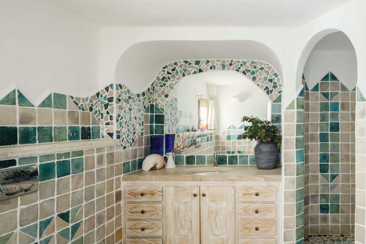 Stylish bathroom featuring patterned green and beige tiles, a wooden vanity with multiple drawers, and an archway design that adds a touch of elegance. Natural light illuminates the space, highlighting the decorative details and a potted plant on the countertop. | Only on SopranoVillas