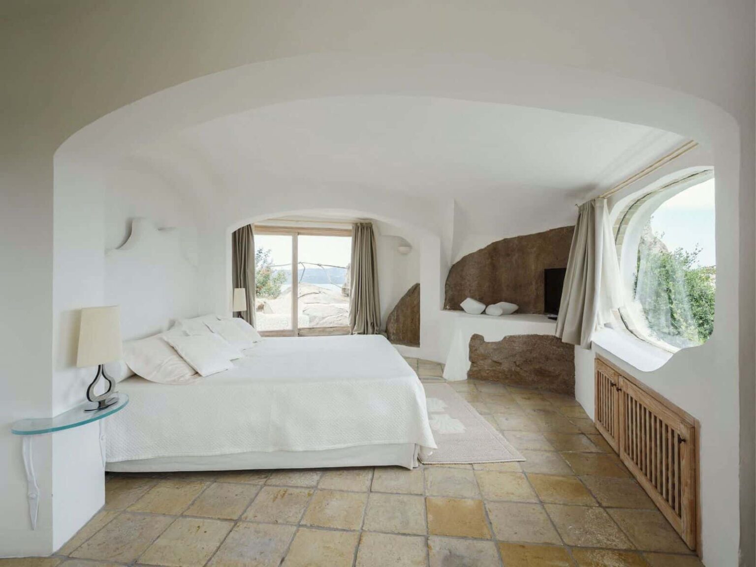 Spacious, modern bedroom with a large window offering scenic views, featuring a comfortable king-size bed, minimalist decor, and natural stone accents. | Only on SopranoVillas