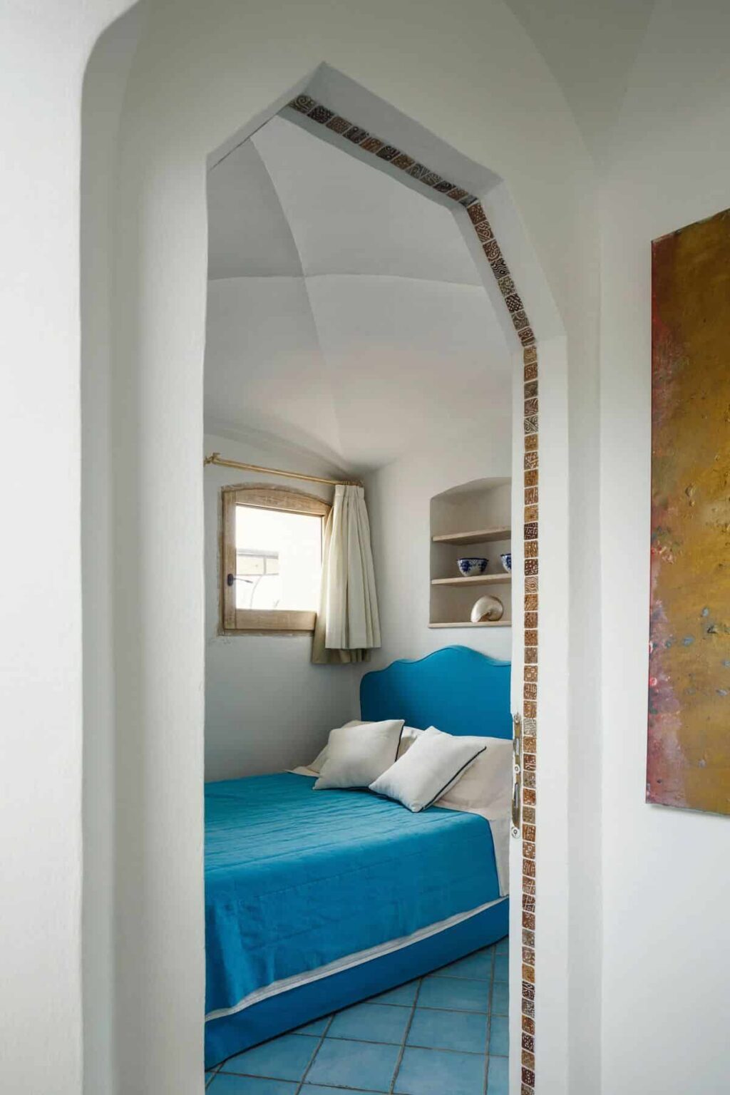 Cozy bedroom featuring a blue bedspread and matching headboard, with natural light streaming through a window adorned with white curtains. The room showcases a minimalist design with white walls, tiled flooring, and decorative shelves. An arched doorway adds architectural interest to the space. | Only on SopranoVillas