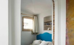Cozy bedroom featuring a blue bedspread and matching headboard, with natural light streaming through a window adorned with white curtains. The room showcases a minimalist design with white walls, tiled flooring, and decorative shelves. An arched doorway adds architectural interest to the space.