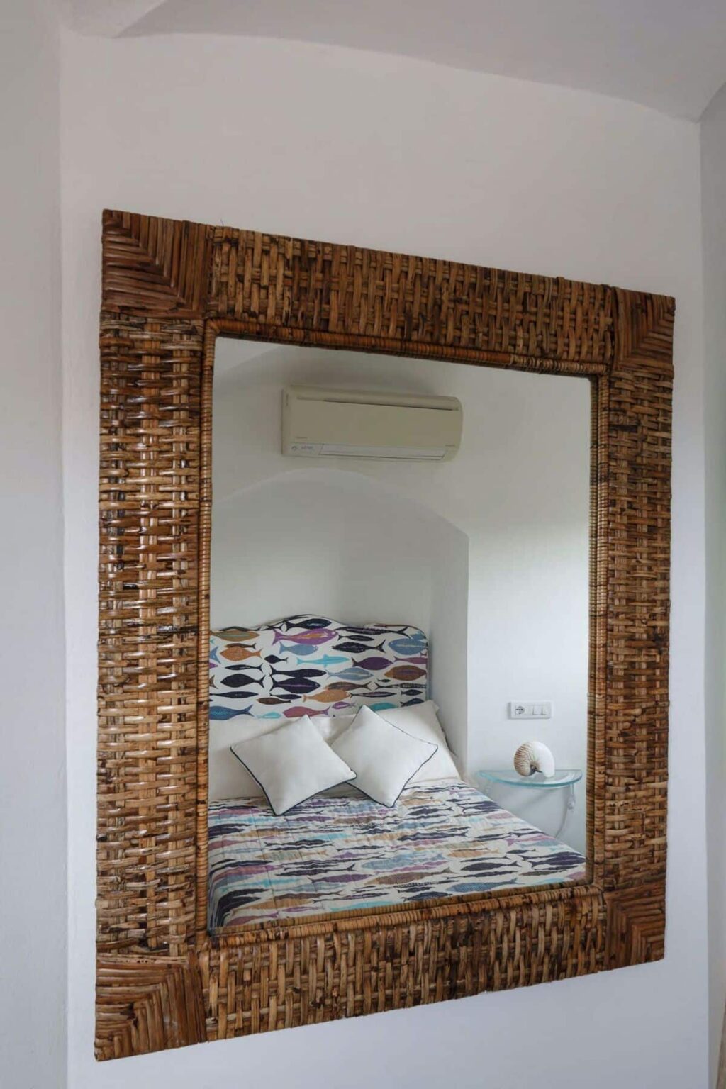 Woven rattan mirror frame reflecting a cozy bedroom with a colorful fish-patterned headboard and decorative pillows, featuring a wall-mounted air conditioner in a bright, minimalist space. | Only on SopranoVillas